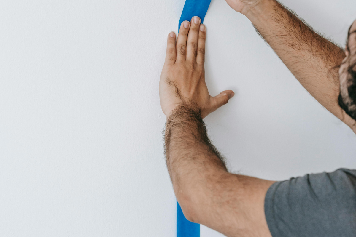 Mão a colar fita-cola azul de pintor ao longo de um bordo de parede branca para delimitar a área de silicone
